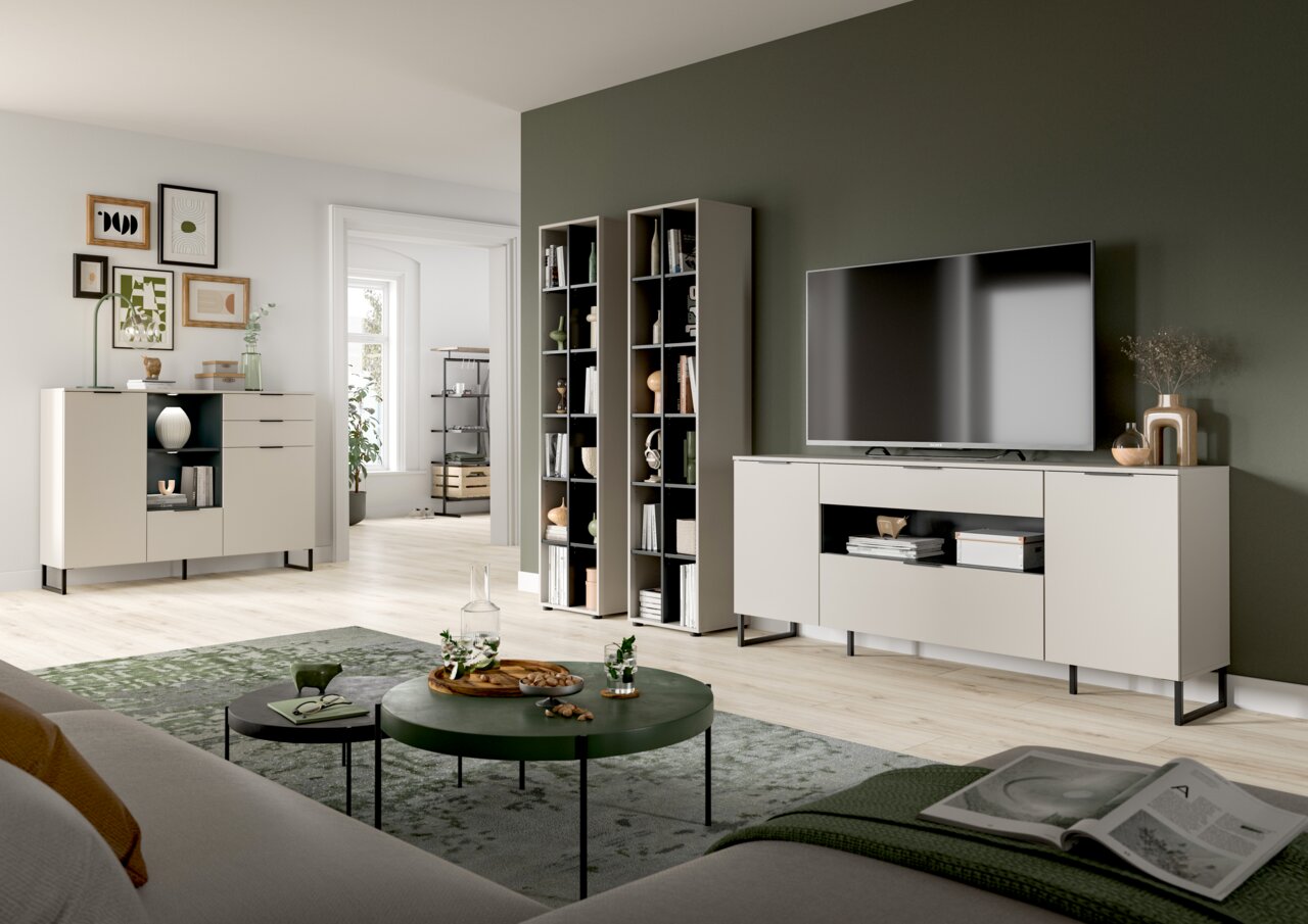 Modernes Wohnzimmer-Set mit hellen Sideboards und hohen Regalen vor olivgrüner Akzentwand, TV auf Lowboard, runde Couchtische auf Teppich; Perspektive: frontale Weitwinkelansicht aus leicht erhöhter Augenhöhe.