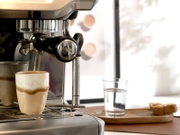Edelstahl-Siebträger-Espressomaschine mit eingespanntem Portafilter, davor ein kleines Glas Latte Macchiato auf der Abtropfschale; Perspektive: Nahaufnahme von vorne links