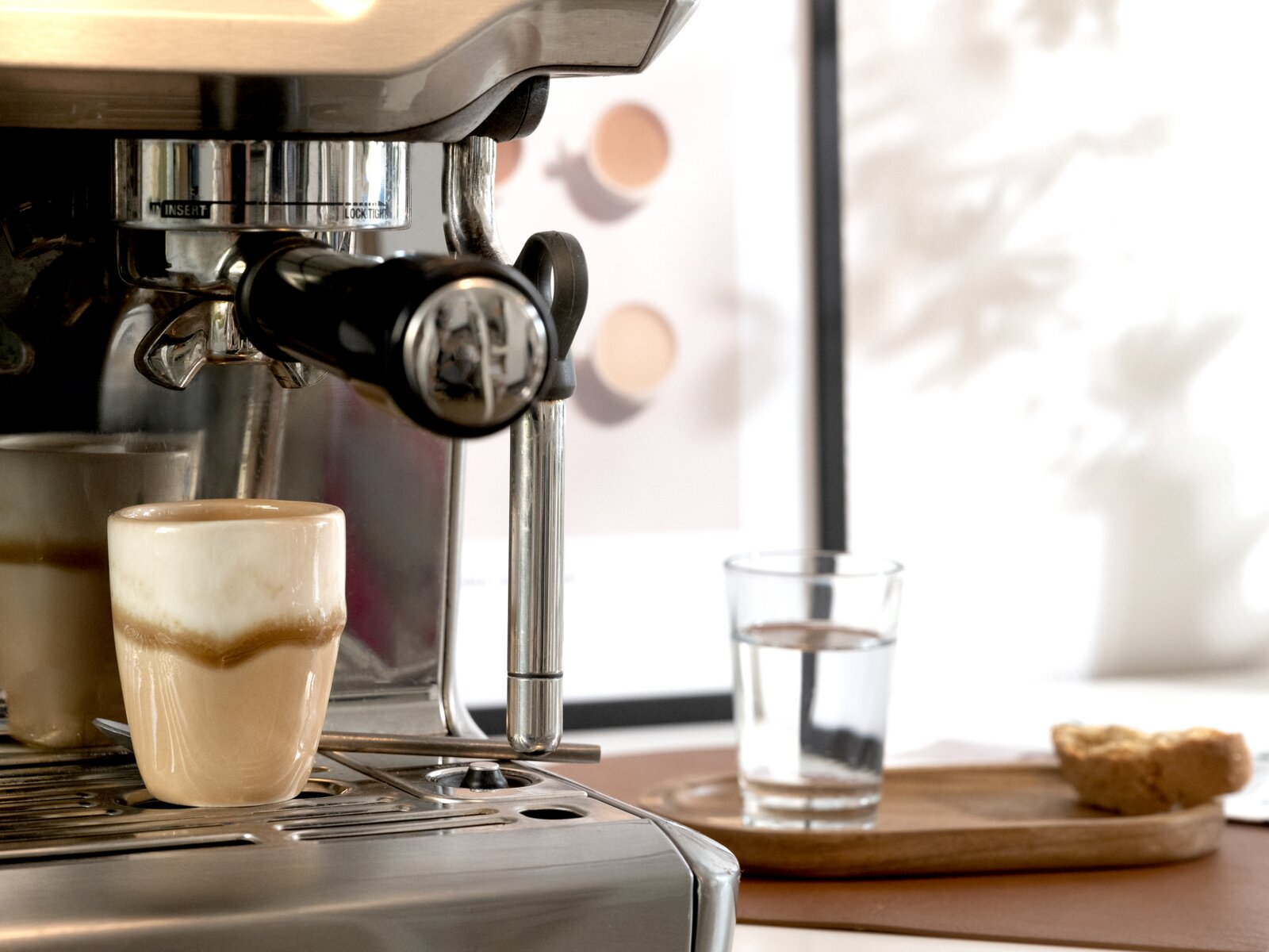 Edelstahl-Siebträger-Espressomaschine mit eingespanntem Portafilter, davor ein kleines Glas Latte Macchiato auf der Abtropfschale; Perspektive: Nahaufnahme von vorne links