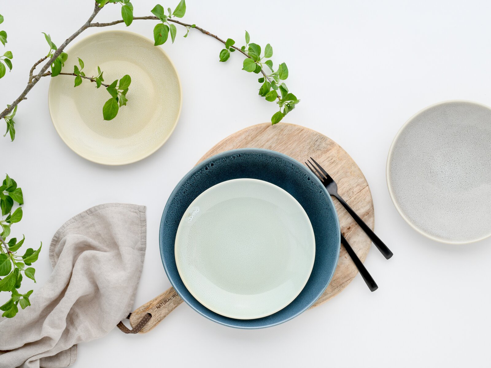 Drei Keramikschalen in Creme, Blau und Grau auf weißem Hintergrund, arrangiert mit Holzbrett, schwarzer Gabel, Leinentuch und grünen Zweigen, Draufsicht von oben