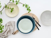Drei Keramikschalen in Creme, Blau und Grau auf weißem Hintergrund, arrangiert mit Holzbrett, schwarzer Gabel, Leinentuch und grünen Zweigen, Draufsicht von oben