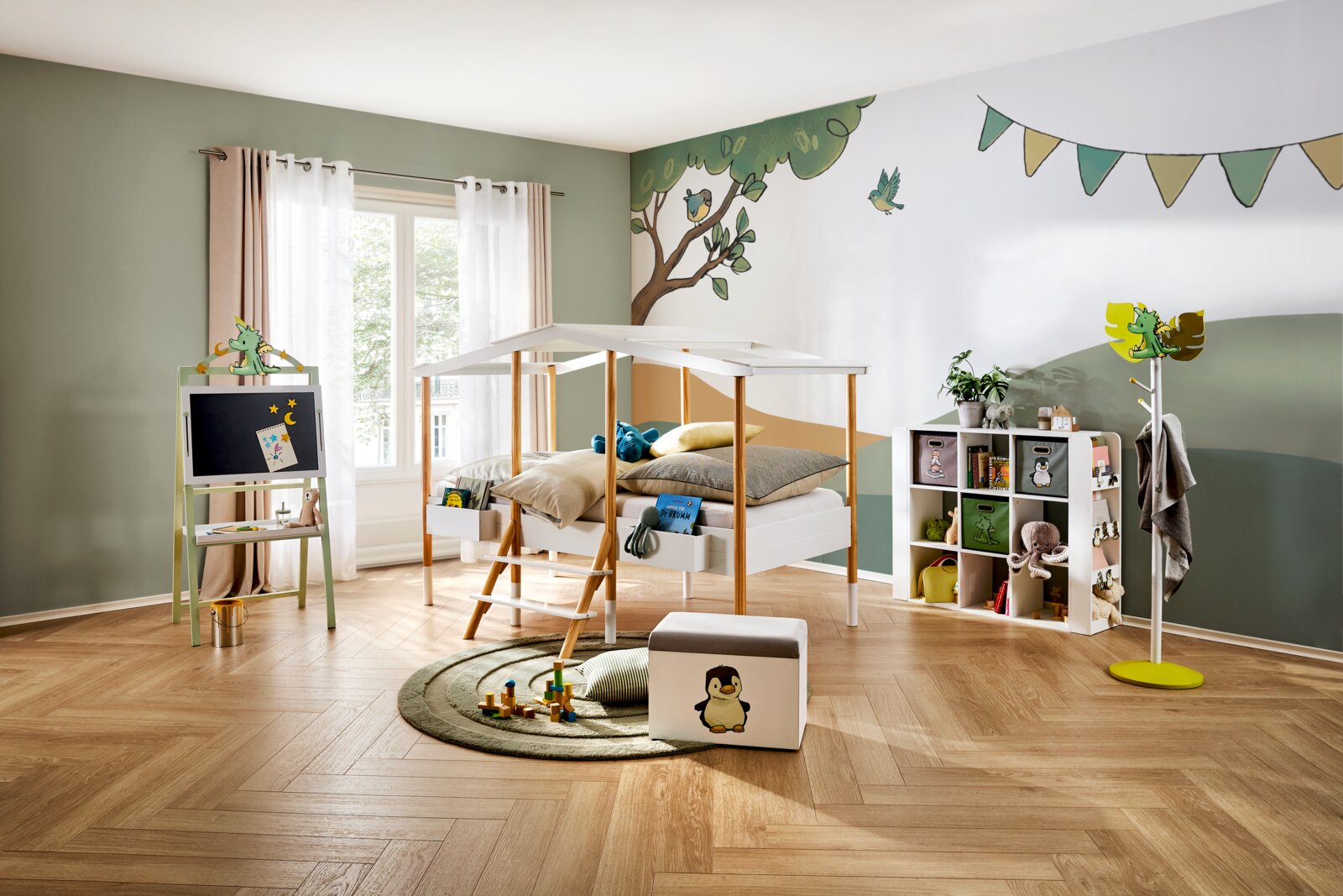 Frontalansicht eines hellen, liebevoll eingerichteten Kinderzimmers mit weißem Hausbett, Maltisch, Spielregal, Garderobe und verspielter Waldtier-Wandgestaltung.