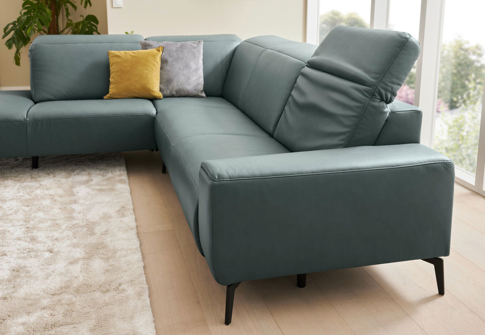 Eckgarnitur in modernem Design mit blauen Polstern, gelbem und grauem Kissen, auf hellem Holzboden und mit Blick auf ein Fenster im Hintergrund.