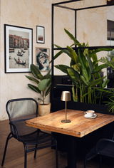 Elegante Akku-Tischleuchte ROXI in einem modernen Café-Setting, auf einem Holztisch neben einer Tasse Kaffee, mit Pflanzen im Hintergrund. Seitenansicht.