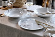 Gedeckter Tisch mit weißen Porzellan-Tellern und Schalen mit italienischem Schriftzug, goldfarbenem Besteck und Weingläsern, fotografiert in schräger Seitenansicht von vorn.
