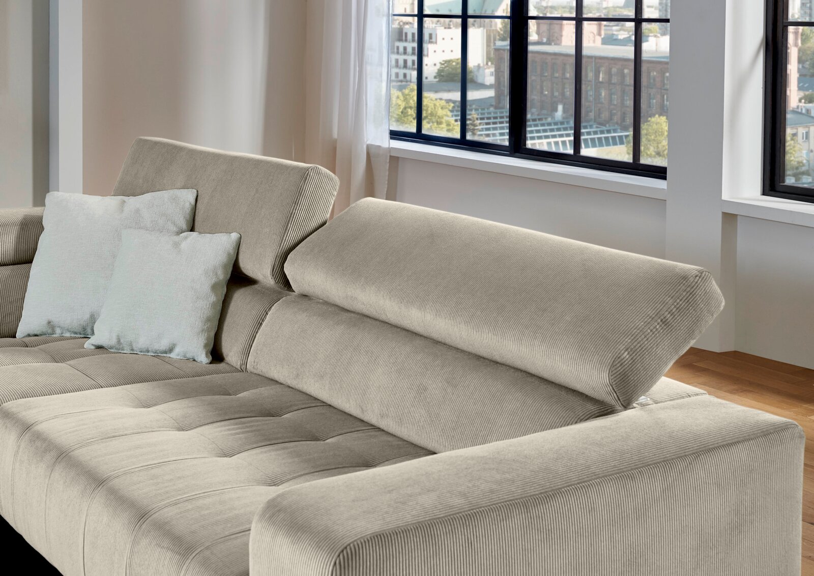 Beiges Cordsofa mit verstellbarer Rückenlehne und zwei hellen Kissen, gezeigt aus schräger Vorderansicht in einem hellen Wohnzimmer mit großen Fenstern.