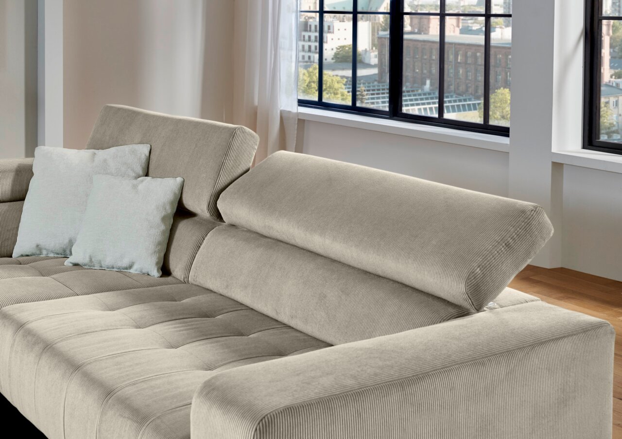 Beiges Cordsofa mit verstellbarer Rückenlehne und zwei hellen Kissen, gezeigt aus schräger Vorderansicht in einem hellen Wohnzimmer mit großen Fenstern.