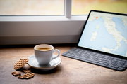 Weiße Espressotasse auf Untertasse mit Waffelkeksen auf einem Holztisch neben einem Tablet mit Kartenansicht am Fenster, aufgenommen aus leicht erhöhter schräger Perspektive
