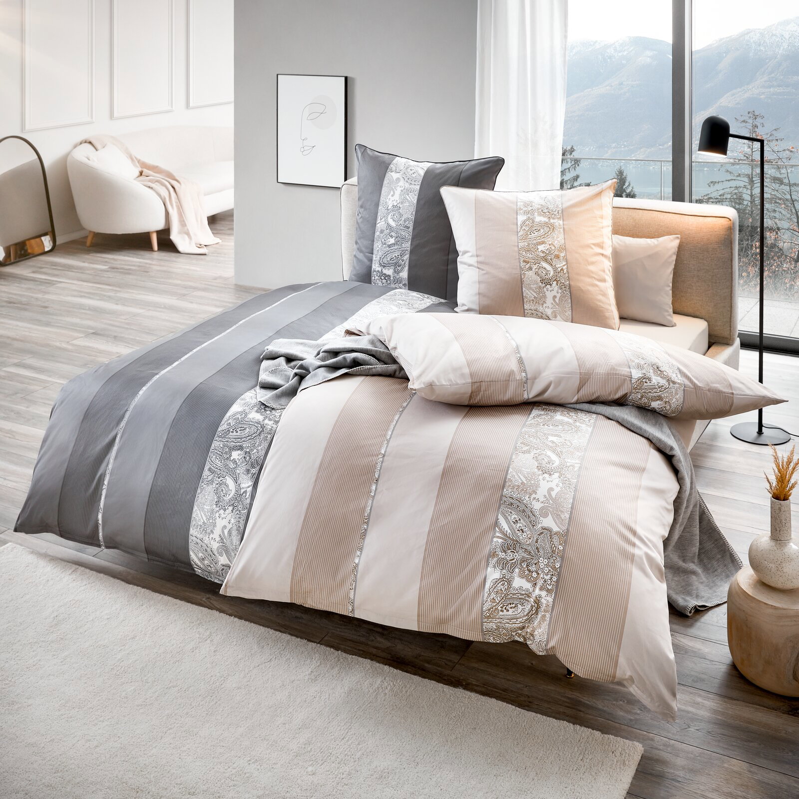 Modernes Bett mit Bettwäsche-Set in Beige, Weiß und Grau mit Streifen- und Paisley-Muster, dekorativen Kissen und Tagesdecke in hellem Schlafzimmer mit großer Fensterfront und Bergblick, Perspektive: schräg von vorne (3/4-Ansicht)