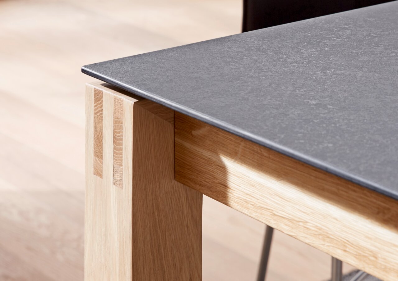 Nahaufnahme eines Esstisches mit einer dunklen Tischplatte und einem hellen Holzgestell, seitliche Perspektive.