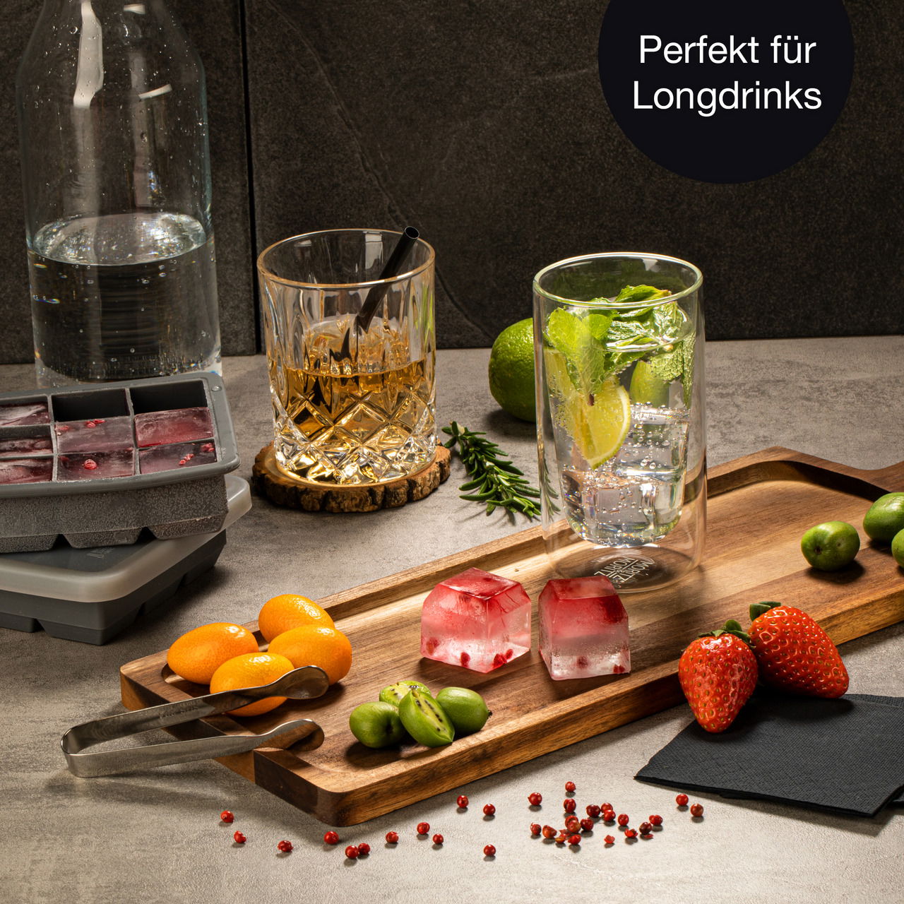 Eiswürfelform mit roten Beeren, präsentiert auf einem Holztablett mit Longdrink-Glas, Früchten und Gewürzen, seitliche Perspektive.