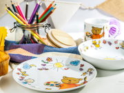 Kindergeschirr-Set aus Porzellan mit Teller, Schüssel und Tasse mit bunten Zeichnungen (Bär, Sonne, Herzen), fotografiert in leichter Schrägansicht von vorne auf einem Tisch mit Buntstiften im Glas im Hintergrund.