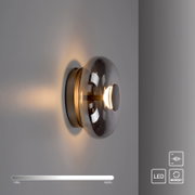Moderne LED-Wandleuchte mit rauchgrauer Glas-Kuppel und Metall-Wandhalterung, eingeschaltet mit warmweißem Licht; seitliche Ansicht in leichter Schrägperspektive von rechts auf einer hellen Wand.