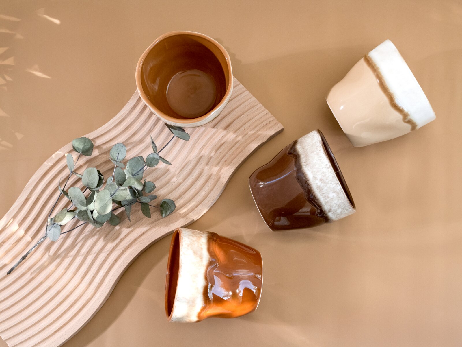 Keramikbecher in Naturtönen (Braun, Beige und Creme) auf Holzbrett mit Eukalyptuszweig, aus schräger Draufsicht fotografiert