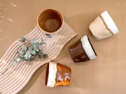 Keramikbecher in Naturtönen (Braun, Beige und Creme) auf Holzbrett mit Eukalyptuszweig, aus schräger Draufsicht fotografiert