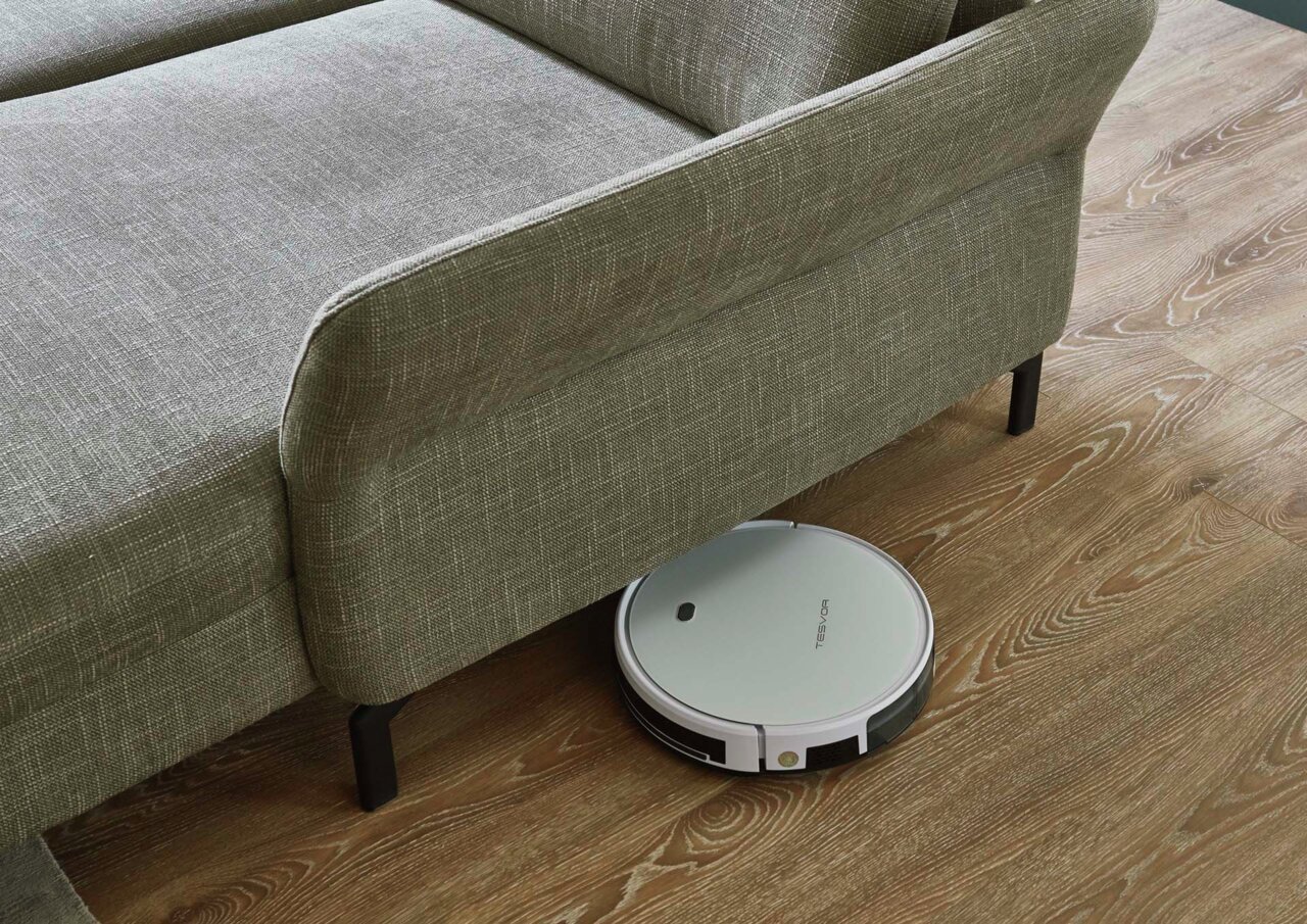 Ein Saugroboter reinigt den Boden unter einer grauen Polsterecke aus der Vogelperspektive.