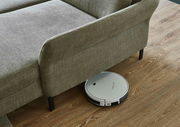 Ein Saugroboter reinigt den Boden unter einer grauen Polsterecke aus der Vogelperspektive.