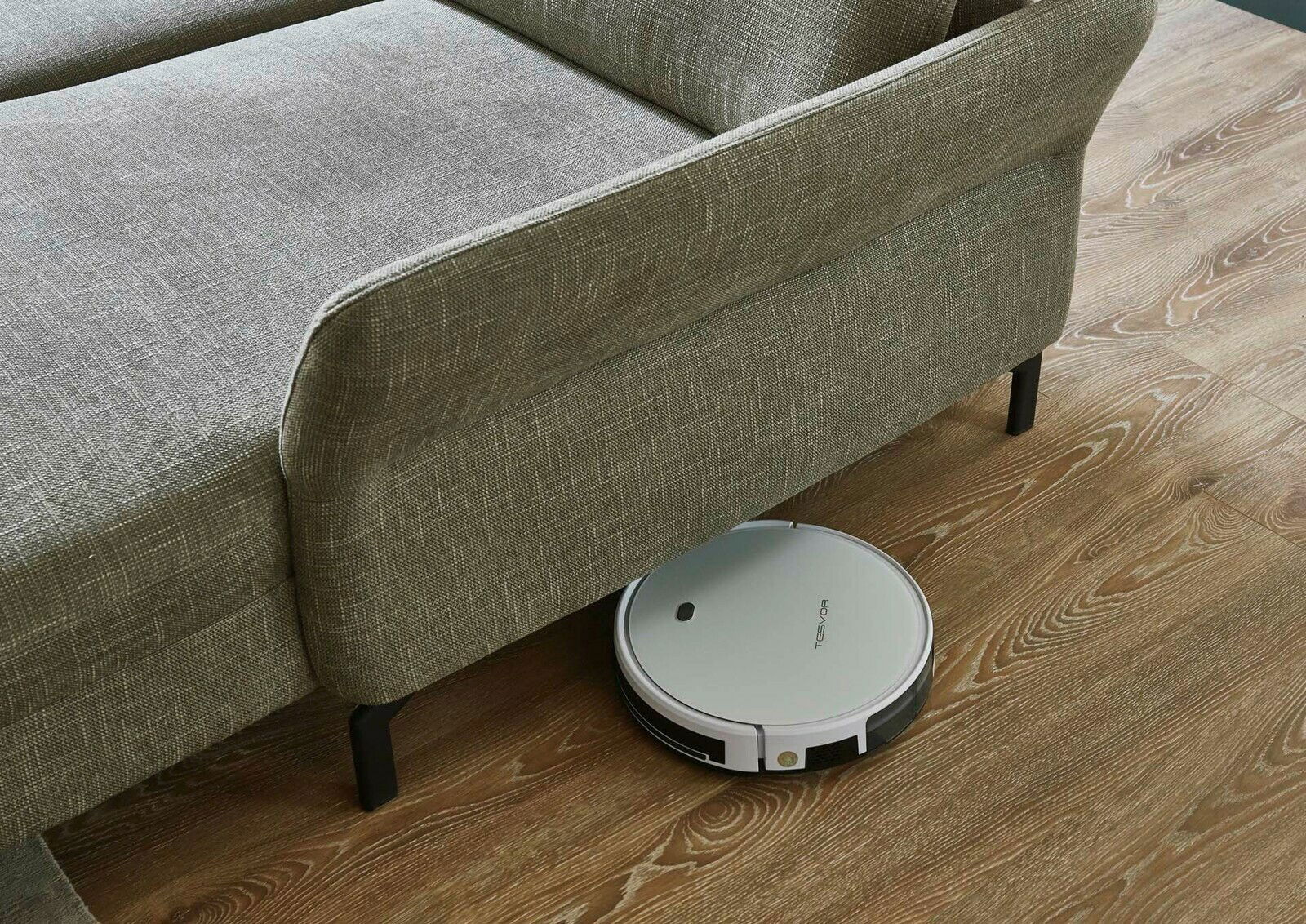 Ein Saugroboter reinigt den Boden unter einer grauen Polsterecke aus der Vogelperspektive.