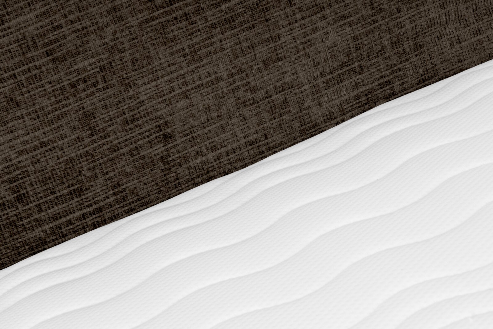 Close-up vanuit schuin bovenaanzicht van een witte matras met subtiel gewatteerd golfpatroon tegen een donkerbruine, gestructureerde bedachterwand.