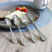 4-teiliges Edelstahlbesteckset mit kindgerechten Gravuren, bestehend aus Löffel, Gabel, Messer und Teelöffel, auf einem Tisch mit Teller und Frühstücksszene, aus der Vogelperspektive fotografiert.