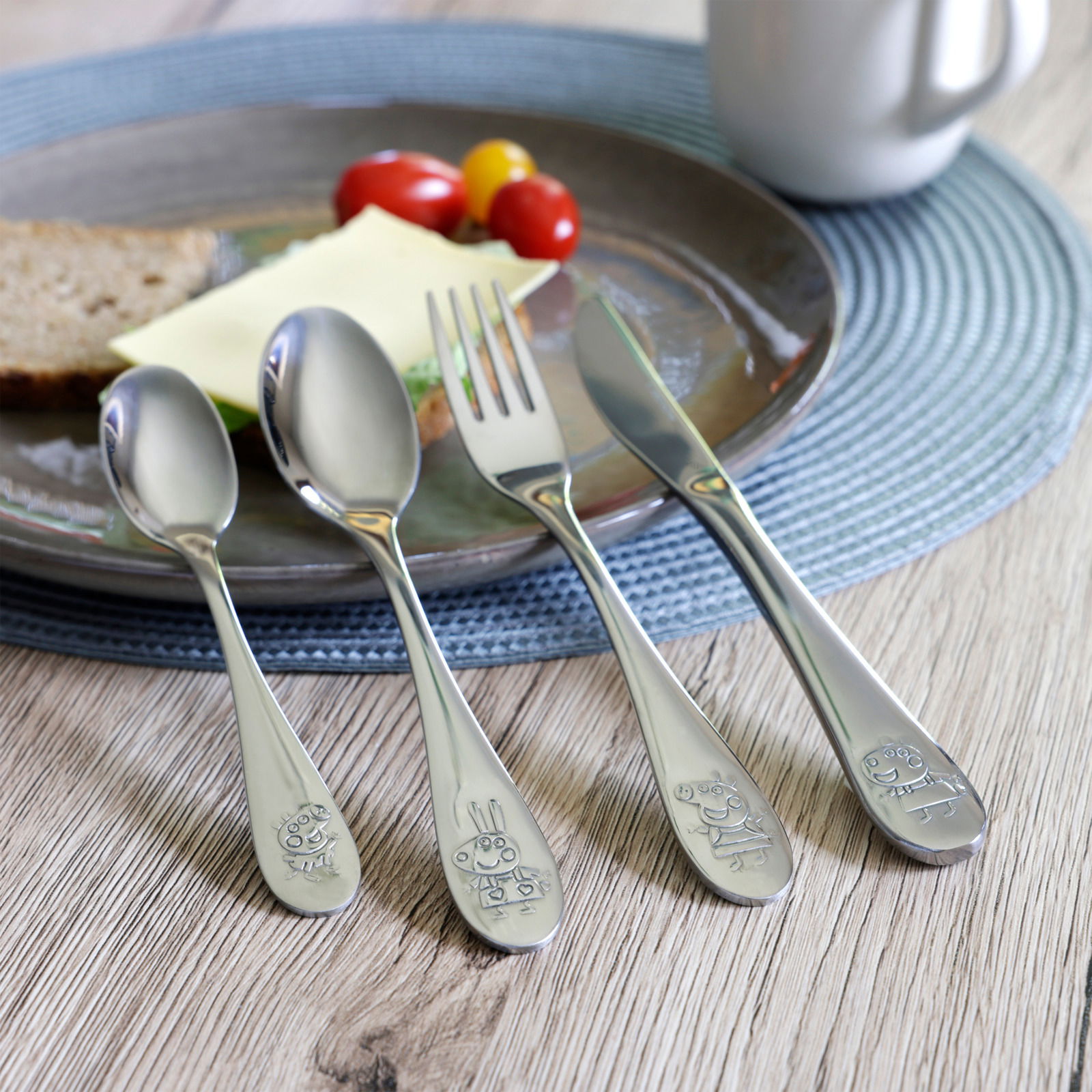 4-teiliges Edelstahlbesteckset mit kindgerechten Gravuren, bestehend aus Löffel, Gabel, Messer und Teelöffel, auf einem Tisch mit Teller und Frühstücksszene, aus der Vogelperspektive fotografiert.