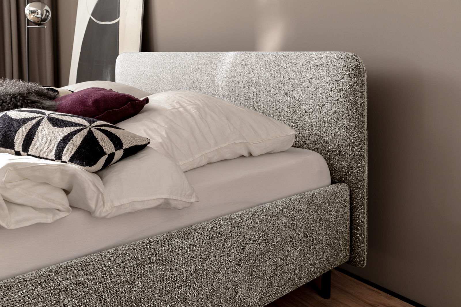 Seitliche Ansicht eines modernen Polsterbetts mit grauem Stoffbezug, dekoriert mit Kissen in verschiedenen Farben und Mustern.