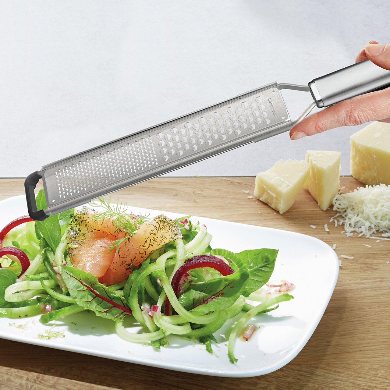 Fein- und Grobreibe PRIMELINE in der Hand gehalten, über einem Teller mit frischem Salat und geriebenem Käse im Hintergrund, seitliche Perspektive.