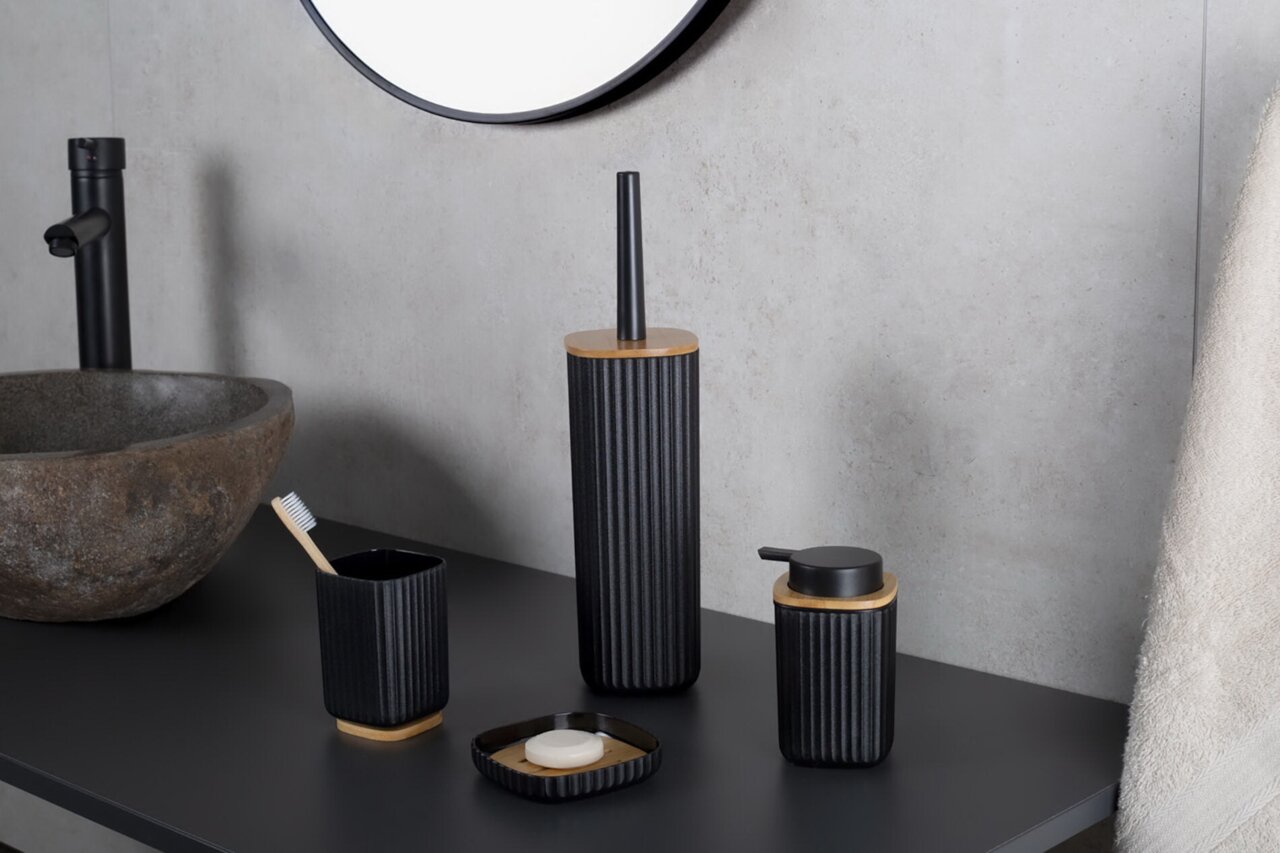 Modernes Bad-Accessoire-Set in Schwarz mit geriffelter Oberfläche und Holzdetails (Toilettenbürste, Seifenspender, Zahnputzbecher und Seifenschale) auf dunkler Waschtischplatte neben Steinwaschbecken, Perspektive: leicht schräg von vorne auf Augenhöhe.