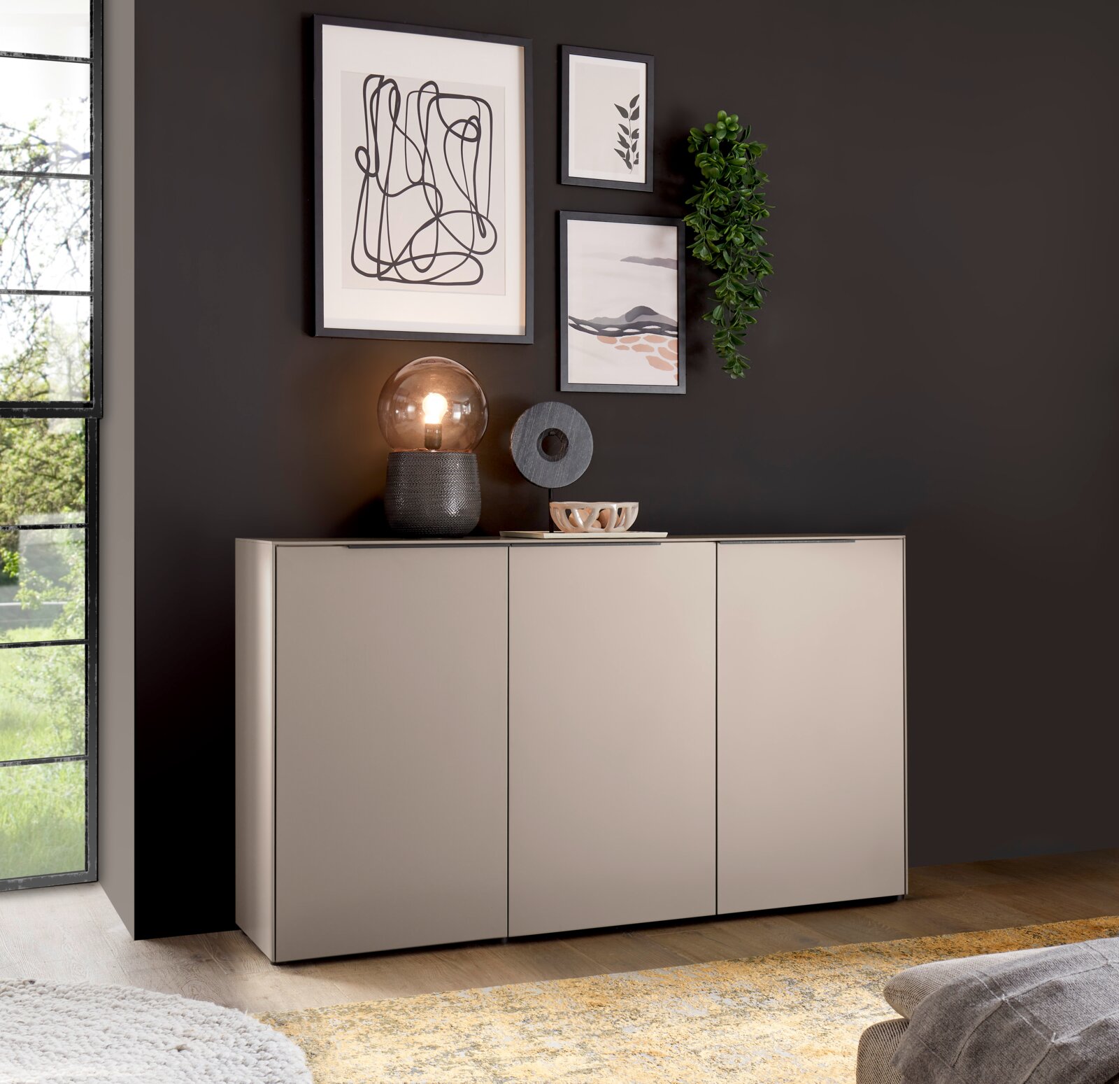 Modernes, hellgraues Sideboard mit drei Fronten vor dunkelgrauer Wand, dekoriert mit Tischlampe, Skulptur, Schale, gerahmten Bildern und hängender Pflanze; Perspektive: frontale Ansicht leicht von links auf Augenhöhe.