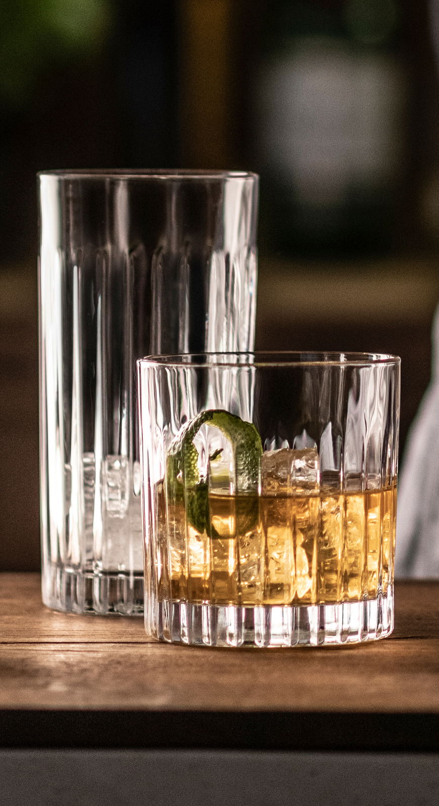 Zwei Whiskygläser auf einem Holztisch, eines gefüllt mit Whisky, Eis und einer Limettenscheibe, aus seitlicher Perspektive.