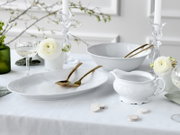 Elegantes weißes Porzellan-Serviergeschirr mit ovaler Platte, großer Schüssel und Sauciere auf festlich gedecktem Tisch mit goldfarbenem Besteck, Glaswaren und weißen Blumen, Perspektive: leicht seitliche Frontansicht auf Tischhöhe.