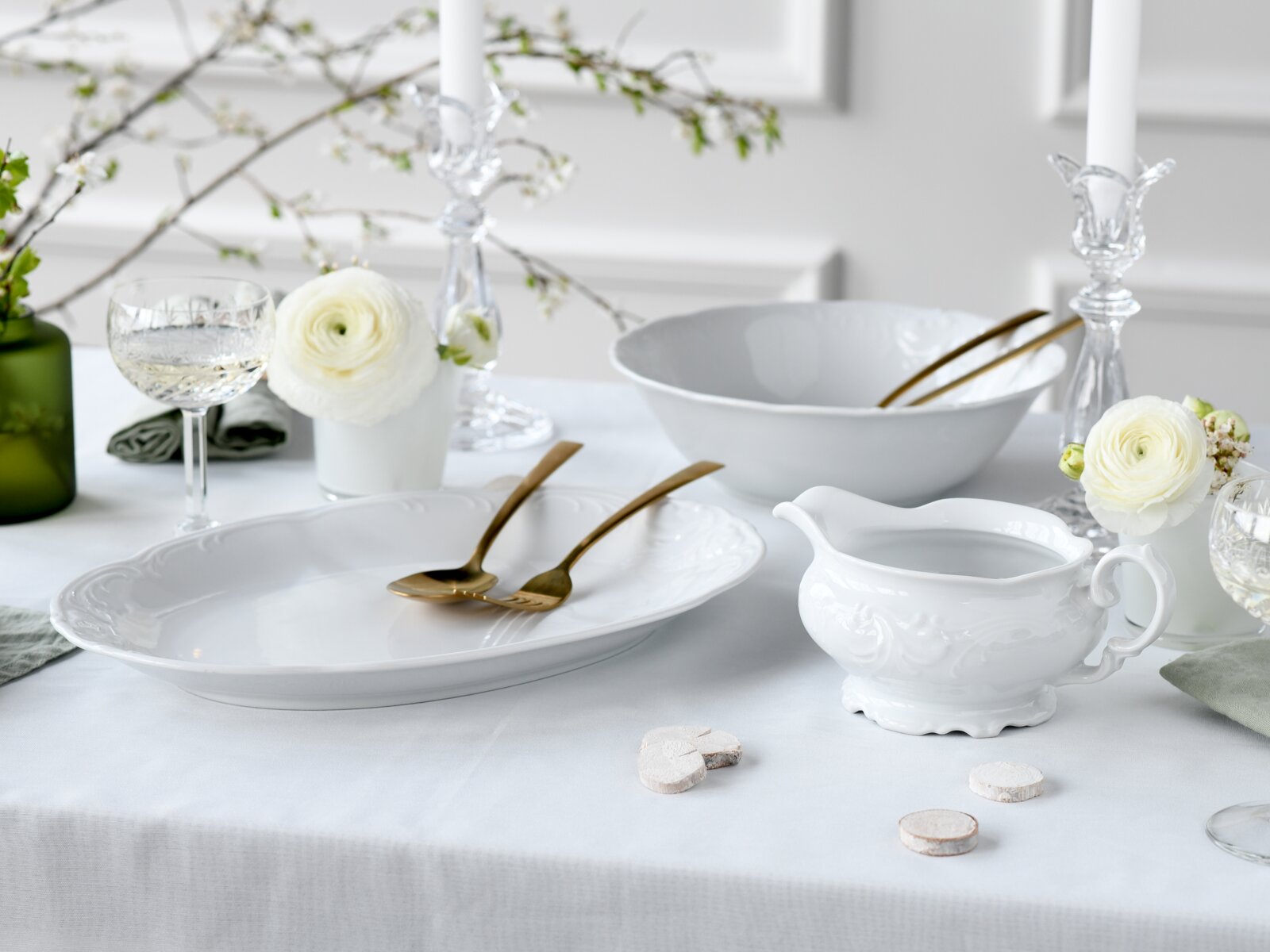 Elegantes weißes Porzellan-Serviergeschirr mit ovaler Platte, großer Schüssel und Sauciere auf festlich gedecktem Tisch mit goldfarbenem Besteck, Glaswaren und weißen Blumen, Perspektive: leicht seitliche Frontansicht auf Tischhöhe.