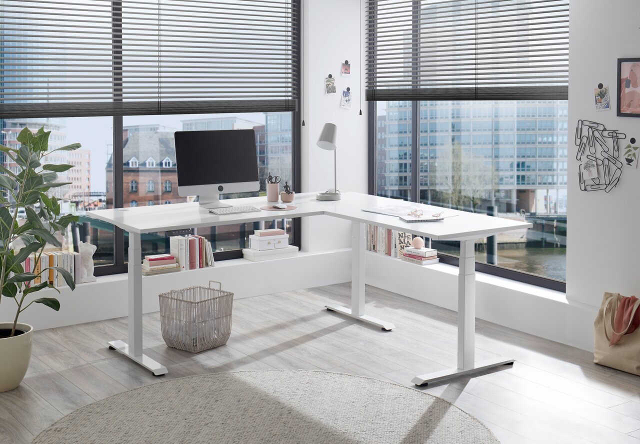 Modern wit elektrisch hoekbureau in een licht kantoor met grote ramen, gezien vanaf de zijkant.
