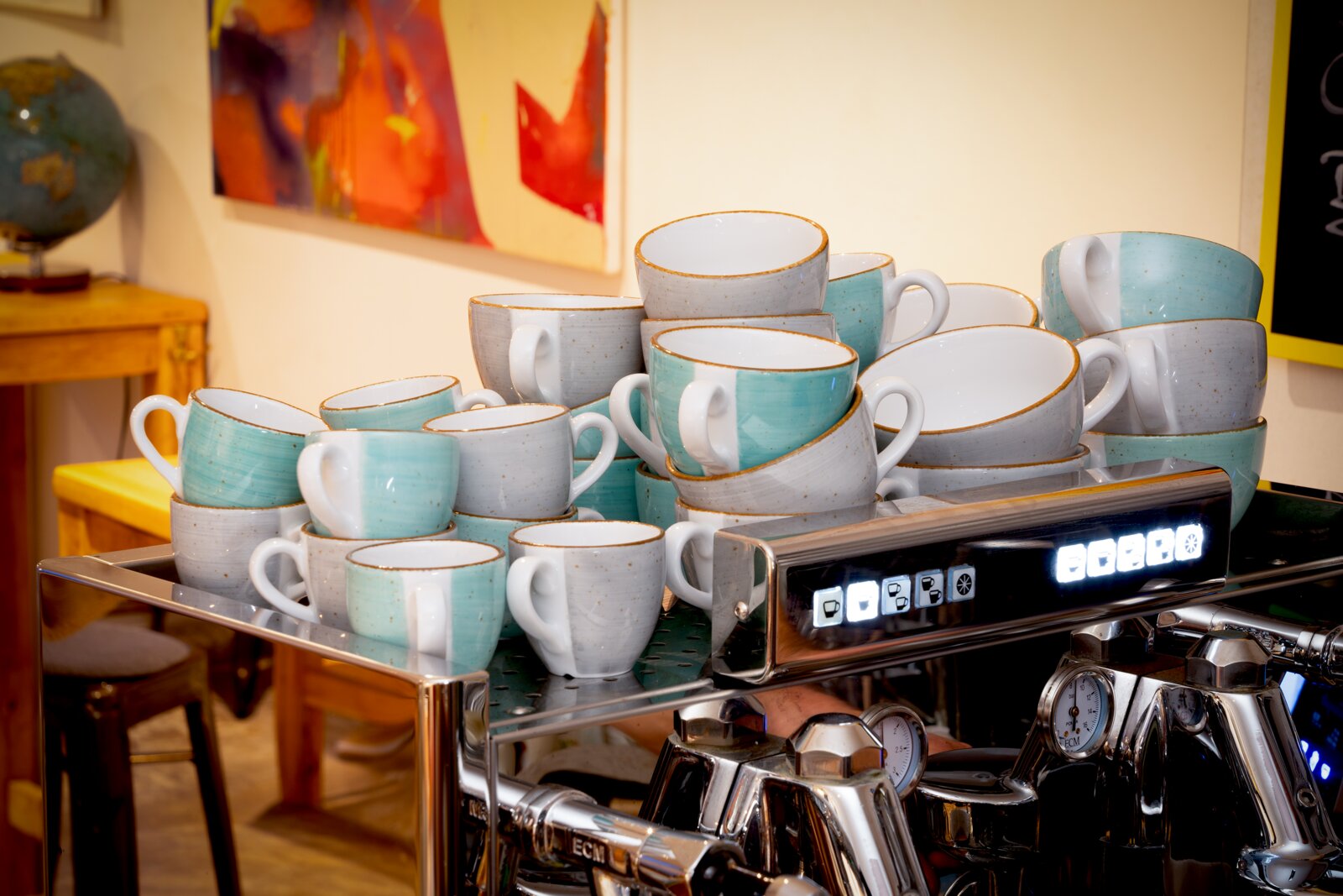 Stapel aus türkis- und grauweißen Keramik-Kaffeetassen mit braunem Rand auf der Oberseite einer Espressomaschine in einem Café, Frontansicht aus leicht erhöhter Perspektive.