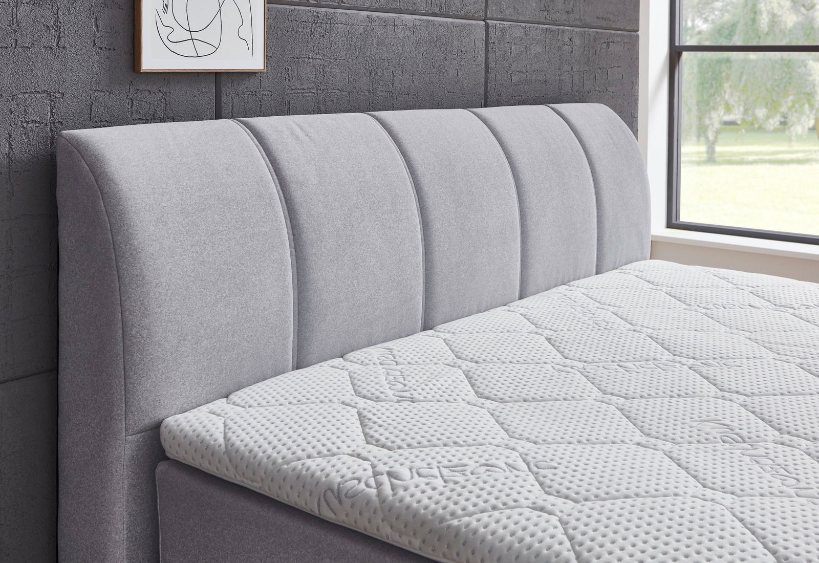 Seitliche Ansicht eines grauen Boxspringbetts mit gepolstertem Kopfteil und strukturierter Matratze in einem modernen Schlafzimmer.