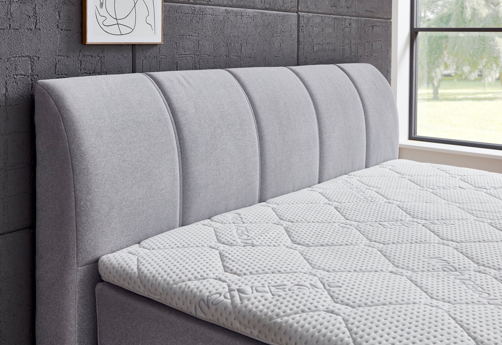 Seitliche Ansicht eines grauen Boxspringbetts mit gepolstertem Kopfteil und strukturierter Matratze in einem modernen Schlafzimmer.