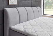 Seitliche Ansicht eines grauen Boxspringbetts mit gepolstertem Kopfteil und strukturierter Matratze in einem modernen Schlafzimmer.