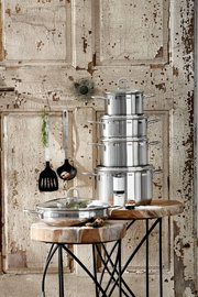 Stapel von glänzenden Edelstahltöpfen auf rustikalem Holztisch vor antiker Holzwand, seitliche Perspektive
