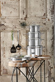 Stapel von glänzenden Edelstahltöpfen auf rustikalem Holztisch vor antiker Holzwand, seitliche Perspektive