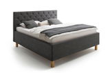 Polsterbett in seitlicher Perspektive mit grauem Kopfteil und Holzfüßen, dekoriert mit Kissen und Decke.
