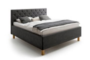 Een gestoffeerd bed in zijaanzicht met een grijs hoofdeinde en houten poten, versierd met kussens en een deken.