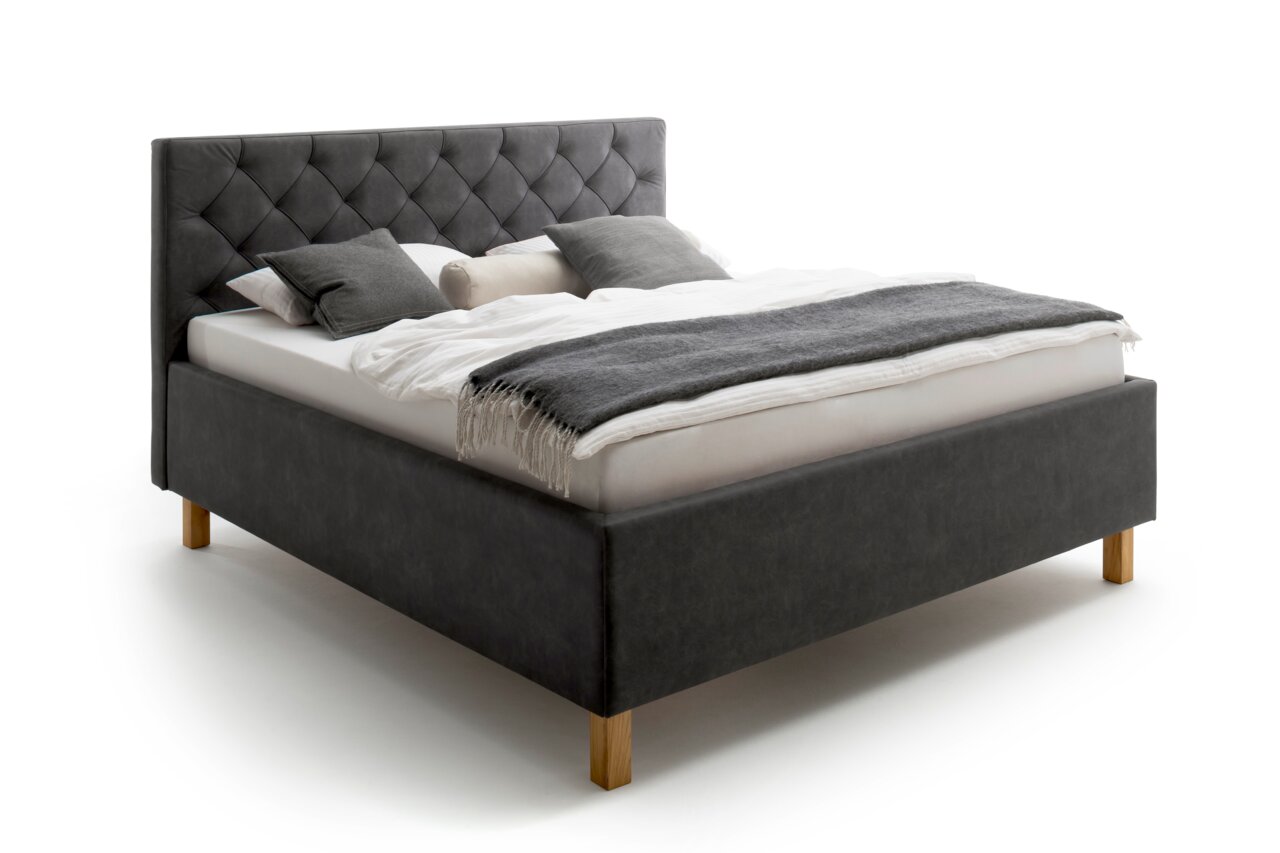 Een gestoffeerd bed in zijaanzicht met een grijs hoofdeinde en houten poten, versierd met kussens en een deken.