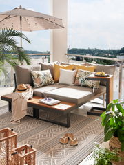 Moderne Gartenlounge mit grauen Kissen und Holzelementen auf einem Balkon mit Blick ins Grüne, seitliche Perspektive.