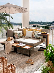 Moderne Gartenlounge mit grauen Kissen und Holzelementen auf einem Balkon mit Blick ins Grüne, seitliche Perspektive.