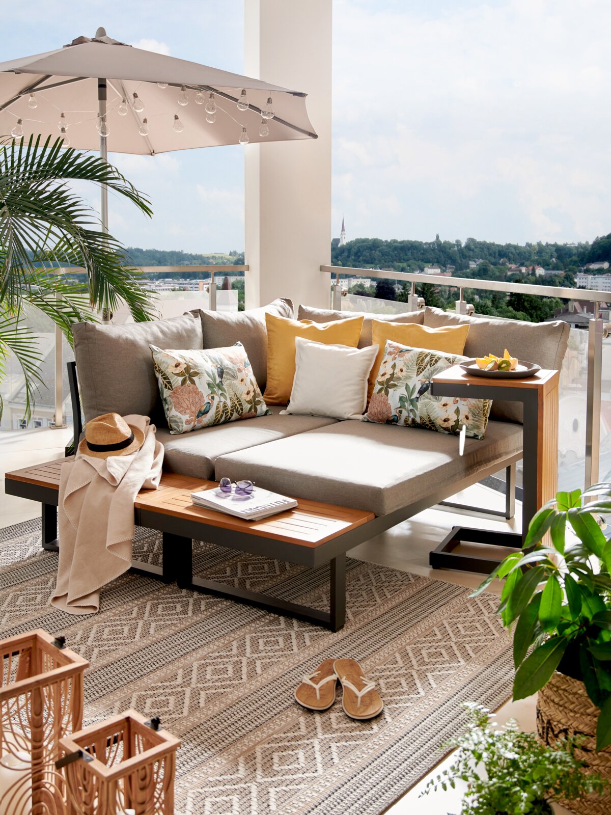 Moderne Gartenlounge mit grauen Kissen und Holzelementen auf einem Balkon mit Blick ins Grüne, seitliche Perspektive.