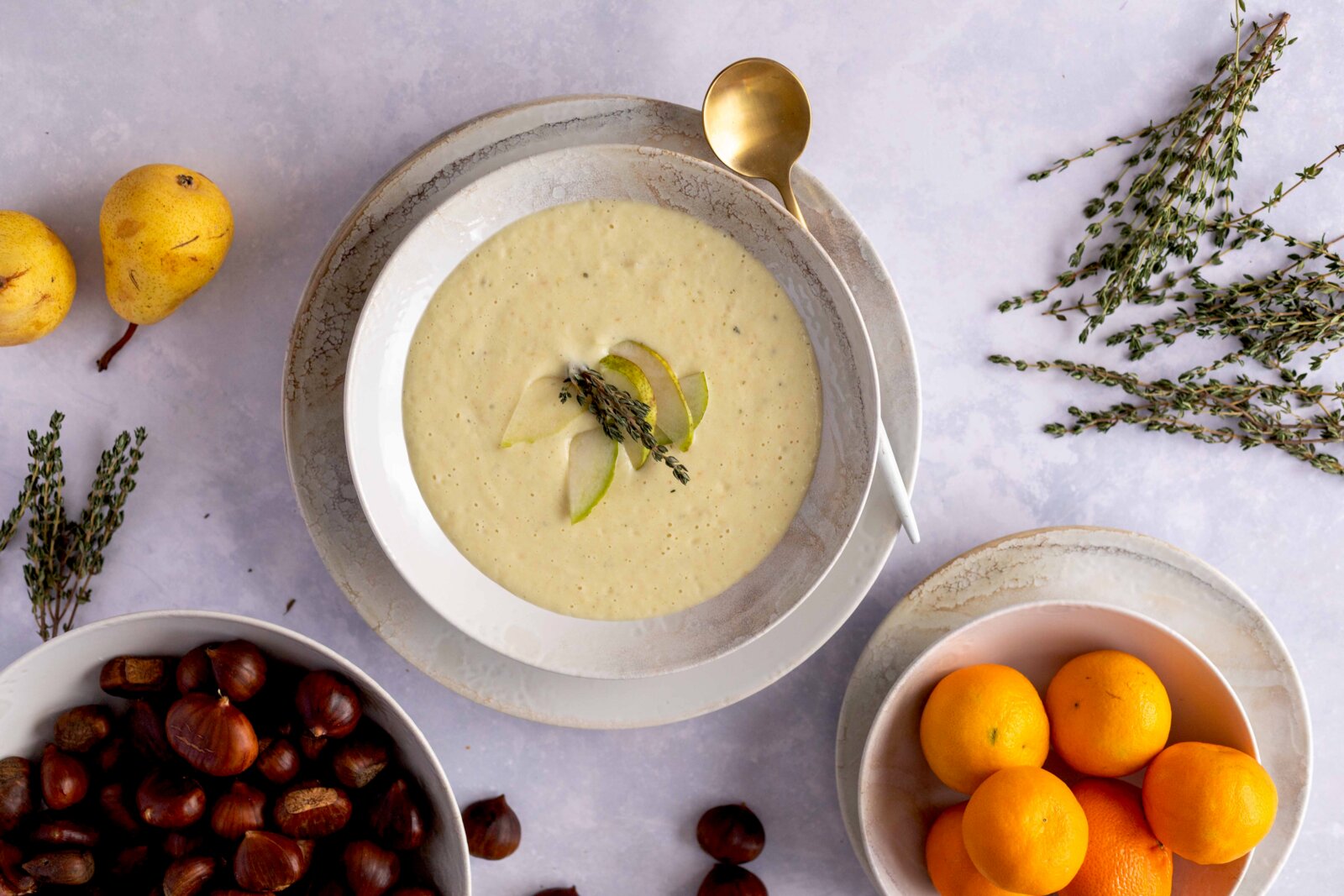 Draufsicht auf eine cremige, helle Suppe in weißer Schale auf Teller, garniert mit Birnenscheiben und Thymian; daneben goldener Löffel, Birnen, Thymianzweige, Kastanien und Orangen auf hellem Hintergrund