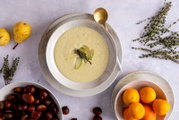 Draufsicht auf eine cremige, helle Suppe in weißer Schale auf Teller, garniert mit Birnenscheiben und Thymian; daneben goldener Löffel, Birnen, Thymianzweige, Kastanien und Orangen auf hellem Hintergrund