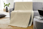 Beige Jacquard Kuscheldecke auf einem grauen Sofa, mit einem Laptop und Kopfhörern auf einem Hocker im Vordergrund, seitliche Perspektive.