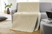 Beige Jacquard Kuscheldecke auf einem grauen Sofa, mit einem Laptop und Kopfhörern auf einem Hocker im Vordergrund, seitliche Perspektive.