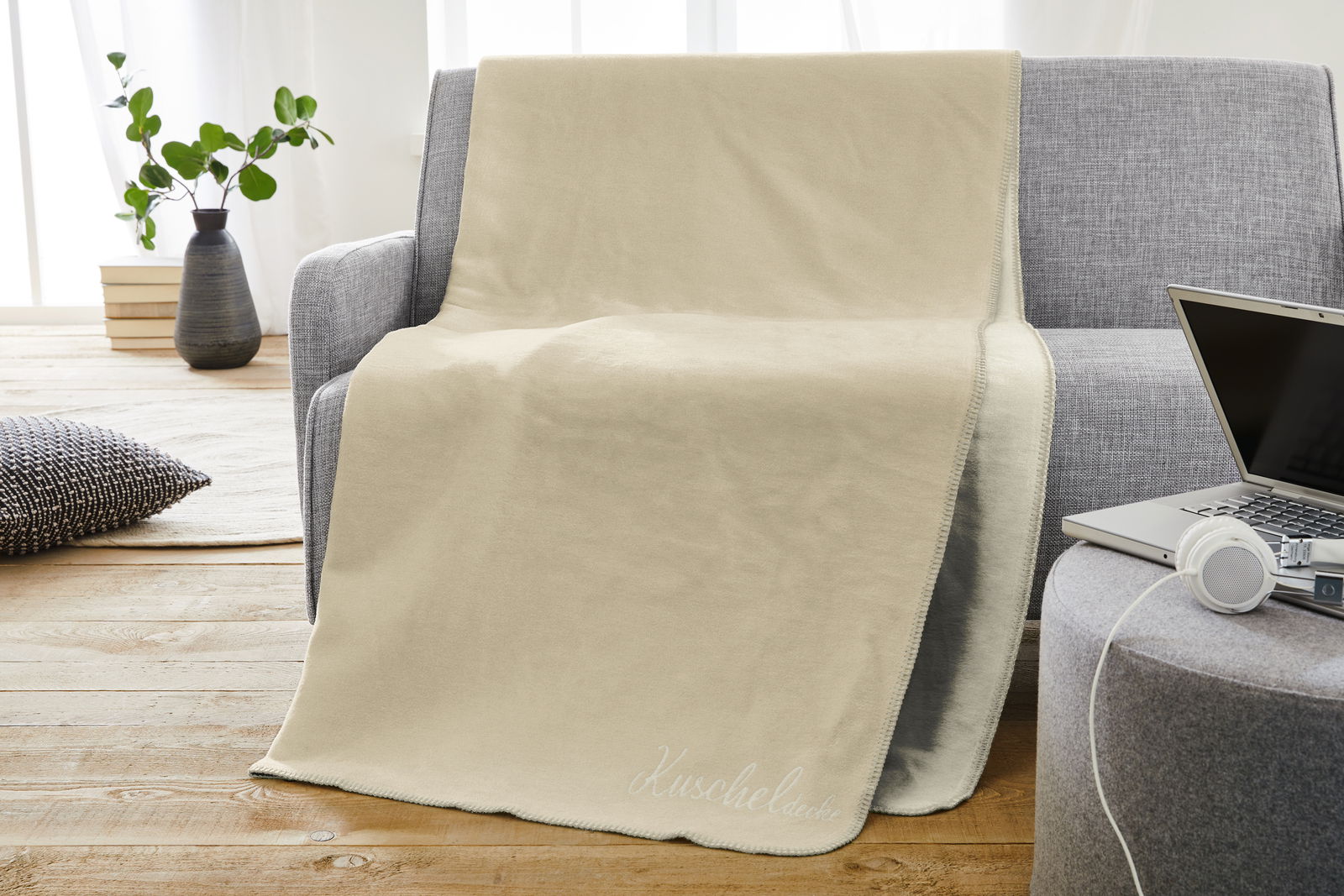 Beige Jacquard Kuscheldecke auf einem grauen Sofa, mit einem Laptop und Kopfhörern auf einem Hocker im Vordergrund, seitliche Perspektive.